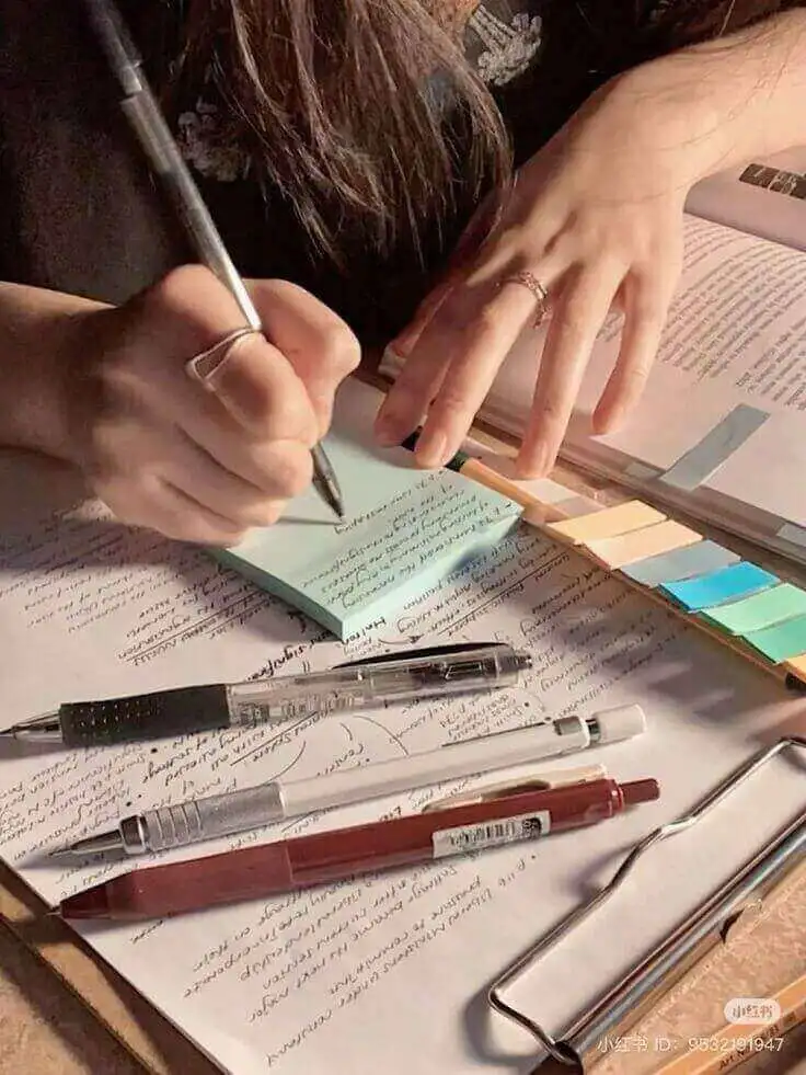 Mãos de um estudante escrevendo um resumo em um bloco de notas adesivo azul claro sobre um livro didático cheio de anotações e marcações, ilustrando o método de estudo ativo.