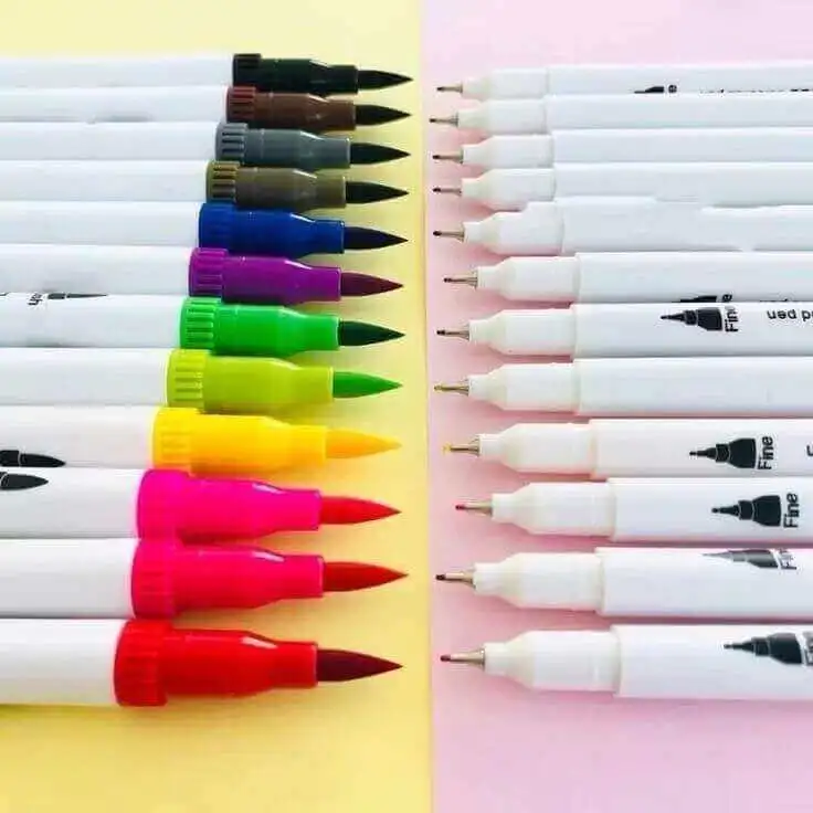 Fileira de canetas coloridas de ponta dupla organizadas simetricamente. À esquerda, aparecem as pontas tipo pincel (brush) e à direita as pontas finas (fineliners), dispostas sobre um fundo de papel amarelo e rosa.