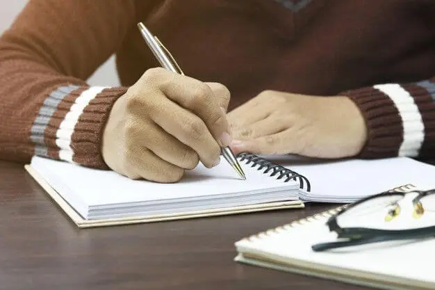 Visão aproximada das mãos de uma pessoa segurando uma caneta metálica sobre um caderno espiral, demonstrando a pegada correta para melhorar caligrafia.